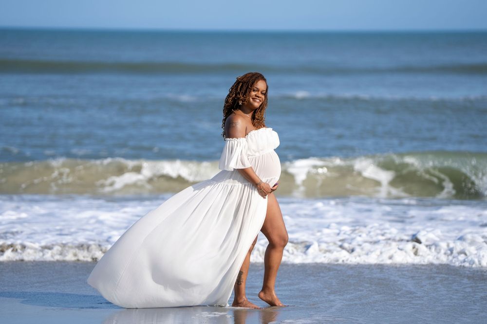A flowing white dress billows in the ocean breeze along a sandy beach with rolling waves in the background.