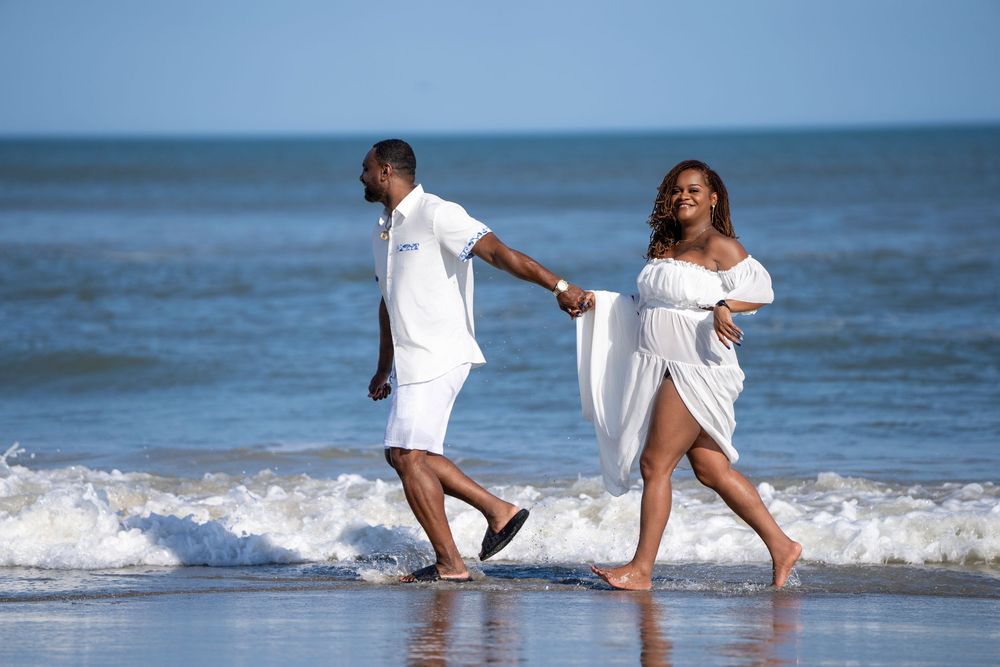 A couple in white clothing walks hand in hand along the beach as ocean waves roll in.