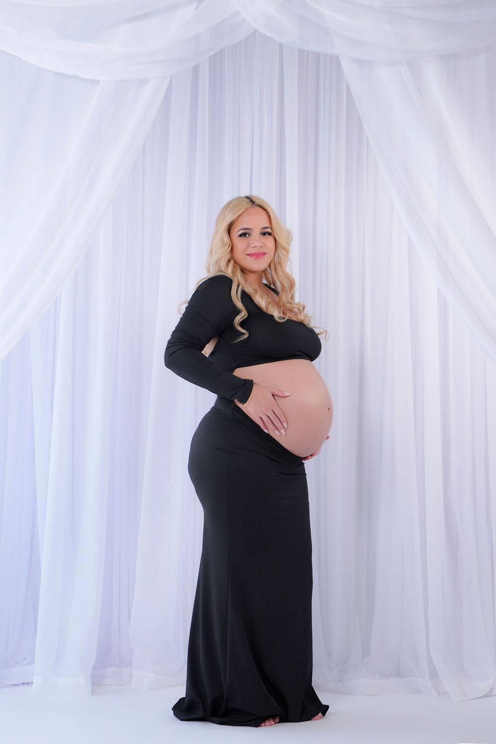 Expectant mother in long black dress poses elegantly against white draped backdrop during maternity photoshoot.