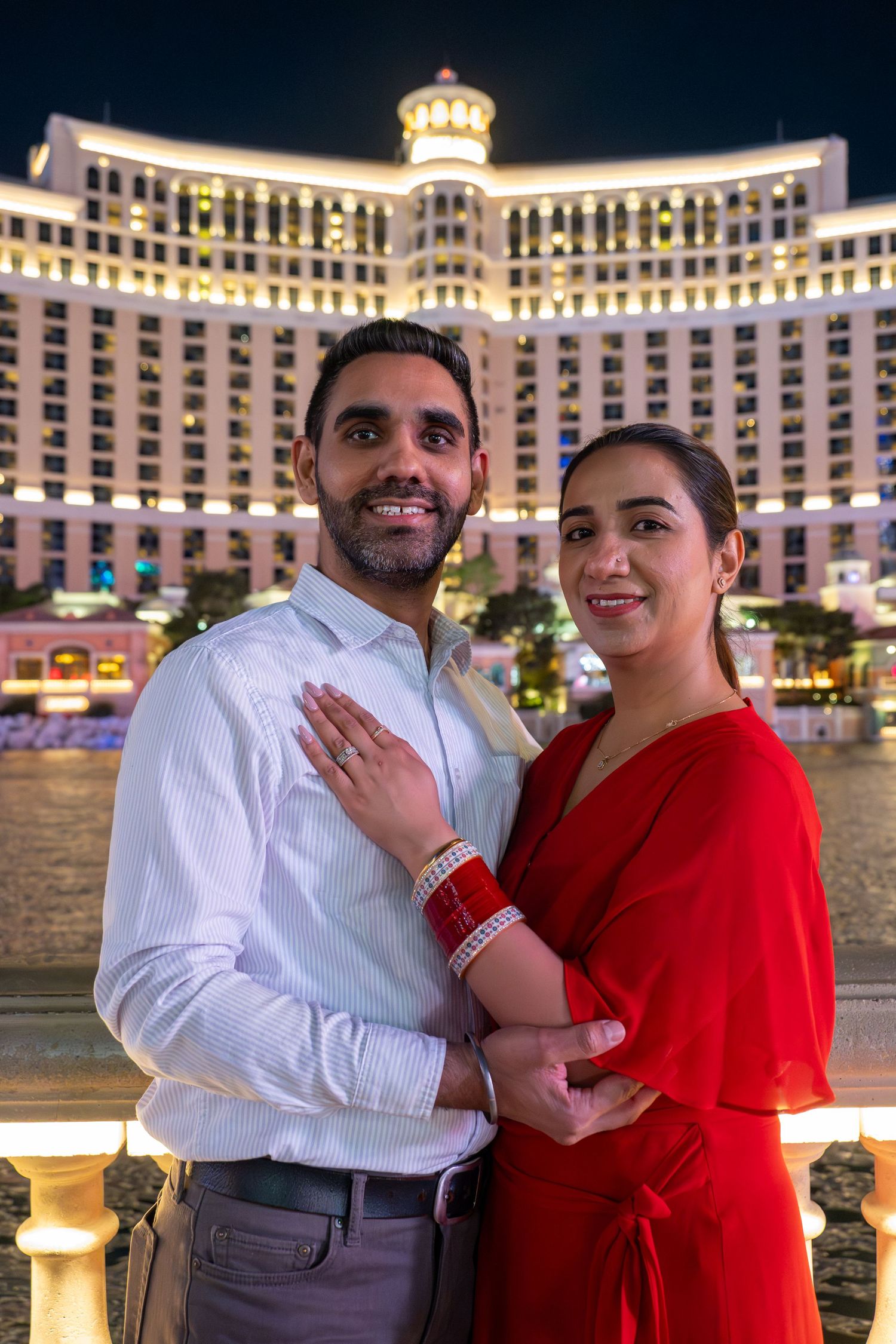 Smiling engaged couple posing at night in front of the illuminated Bellagio Hotel in Las Vegas.