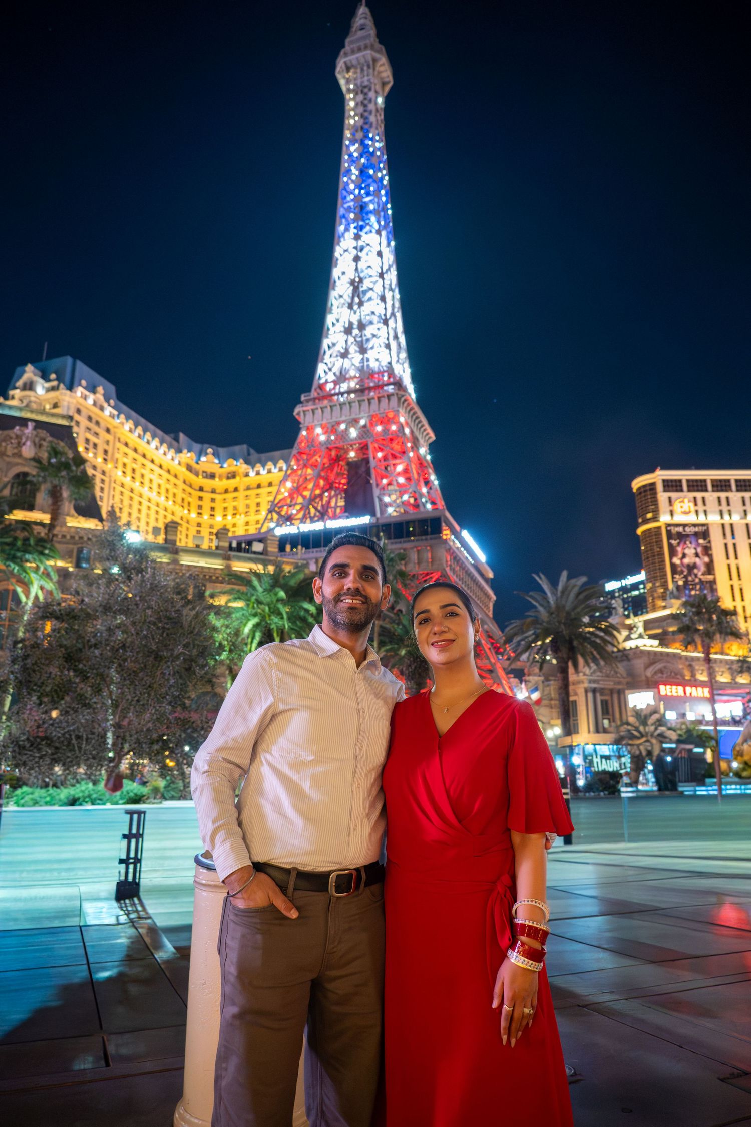 Smiling couple posing in front of the illuminated replica Eiffel Tower at Paris Las Vegas at night.