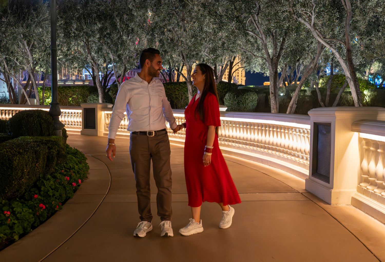 Couple walking hand in hand along a lit pathway surrounded by trees at night.