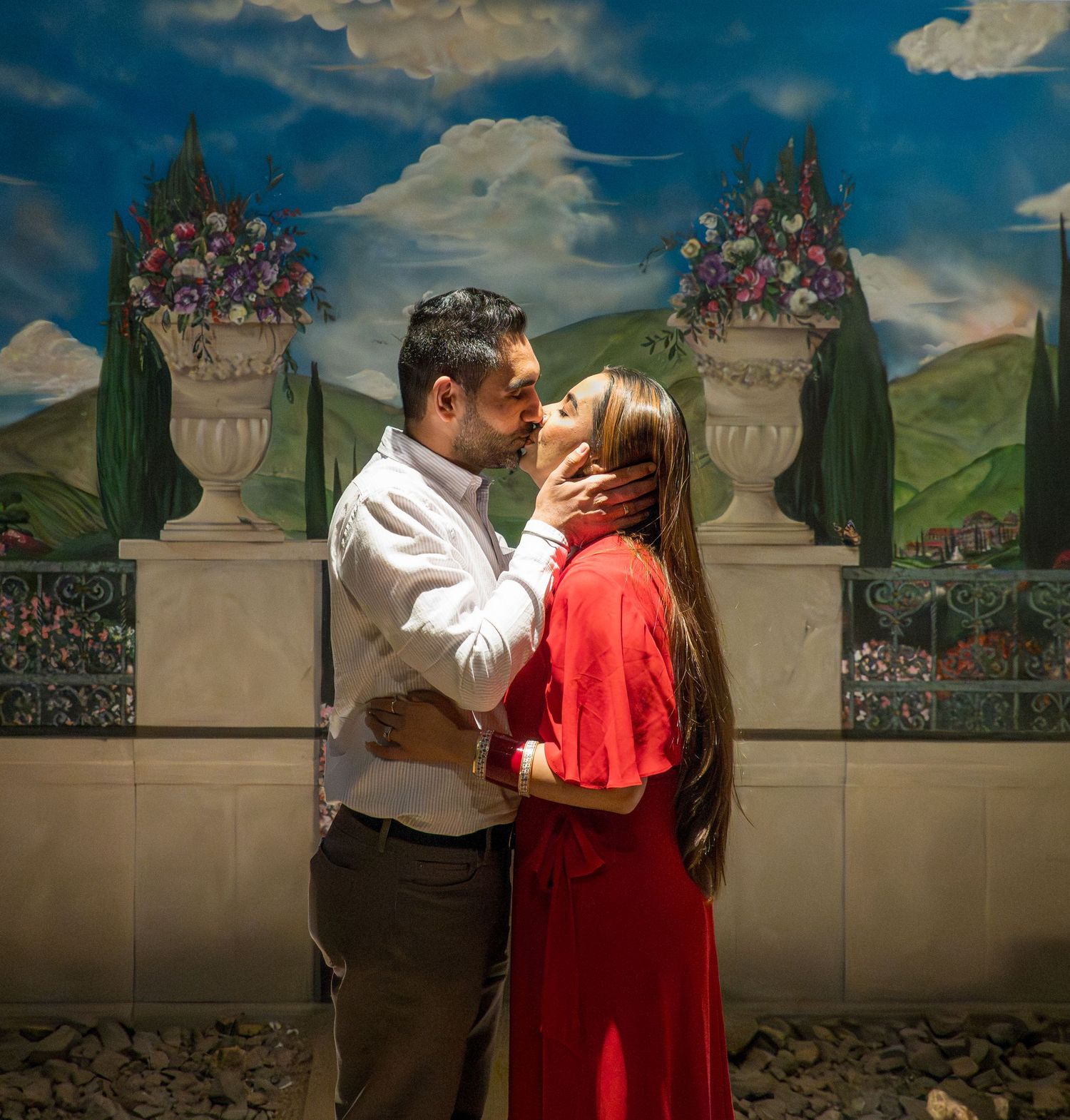 Couple sharing a tender kiss in front of a scenic Italian garden mural with floral urns.