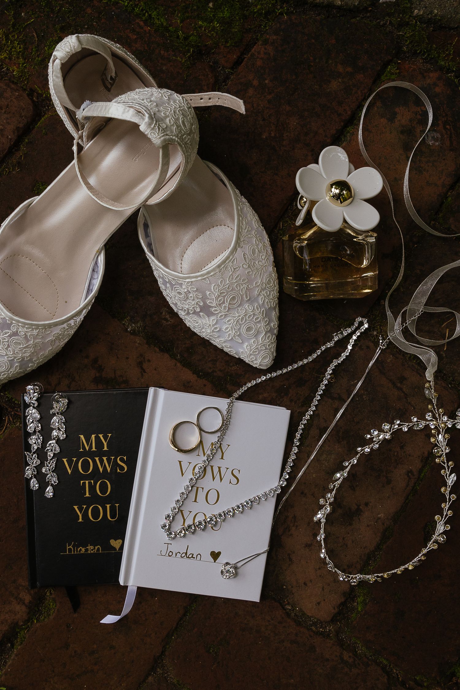 Wedding accessories including white lace shoes, vow books, and jewelry arranged on dark background.