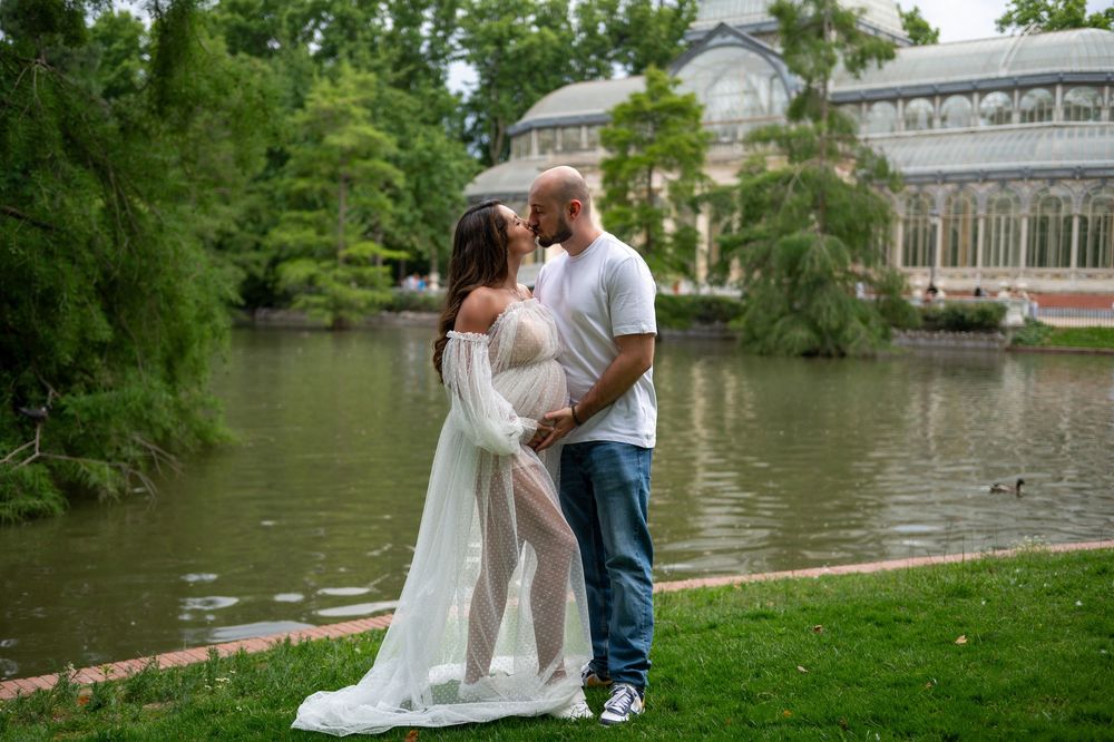 A romantic couple poses for photos near a scenic lake with a glass conservatory building visible in the background.