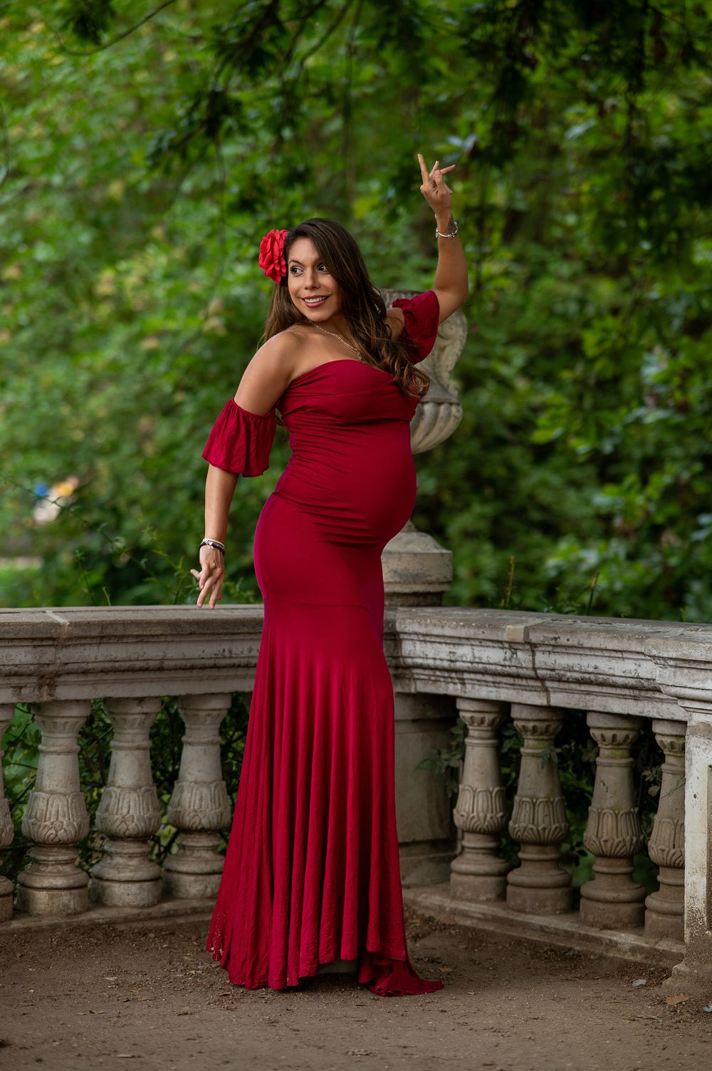 A figure in a flowing red dress poses gracefully against a stone balustrade in a garden setting.