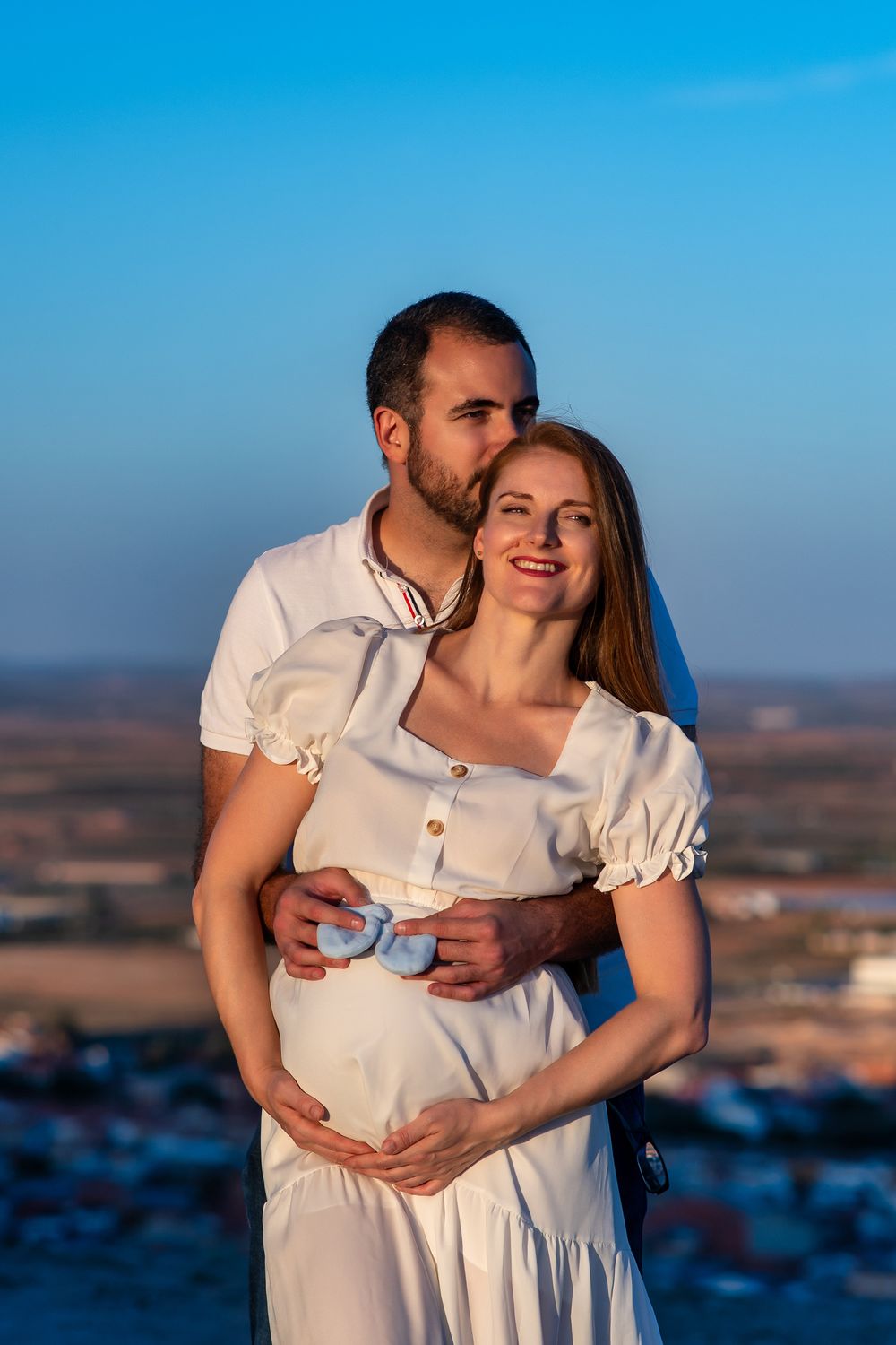A couple in white outfits share an intimate moment against a blue sky backdrop at sunset.