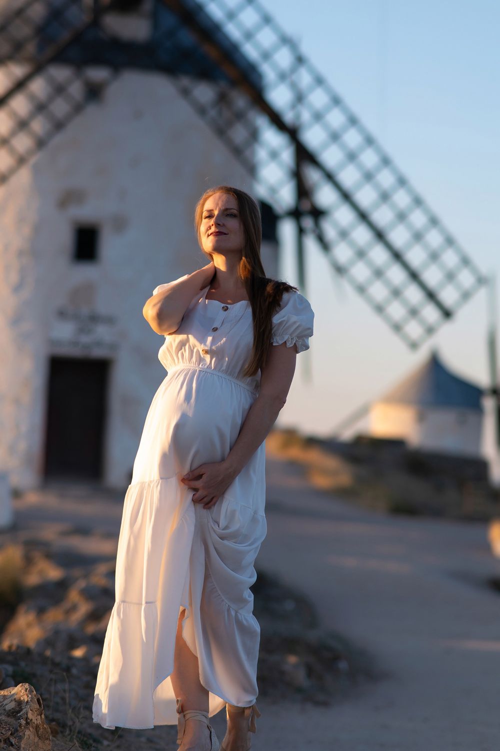 A person in a white dress poses gracefully in front of traditional Spanish windmills at sunset.