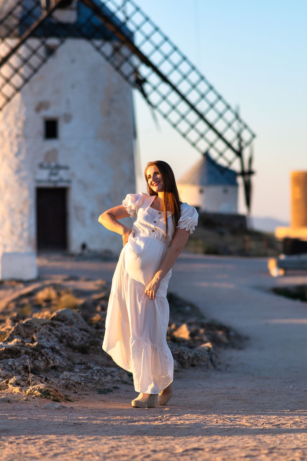 A person in a flowing white dress poses gracefully in front of traditional Spanish windmills at sunset.
