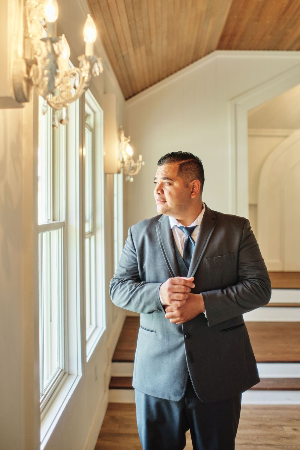 A person in a gray suit adjusts their tie while standing in an elegant room with wooden beams and windows.
