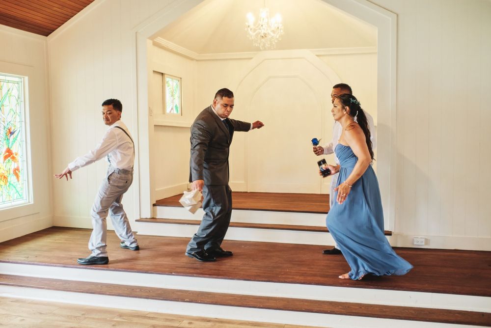 Wedding party members interact playfully in an elegant room with hardwood floors and stained glass windows.