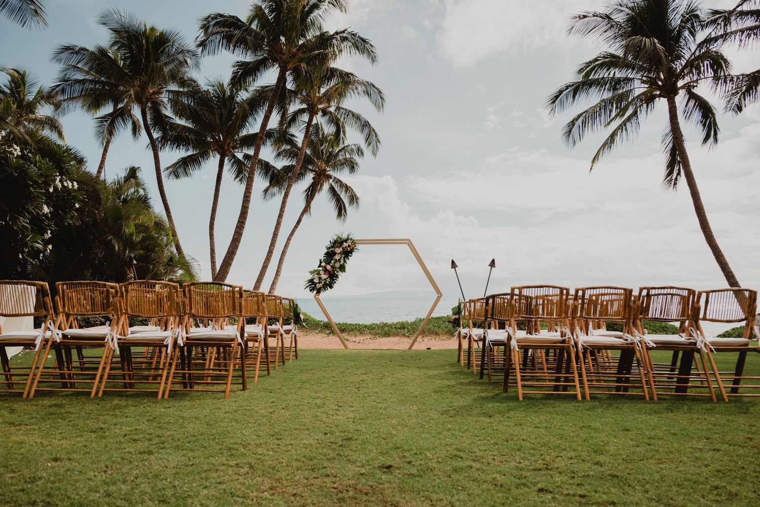 Wooden ceremony chairs set up on a grassy oceanfront lawn with palm trees.