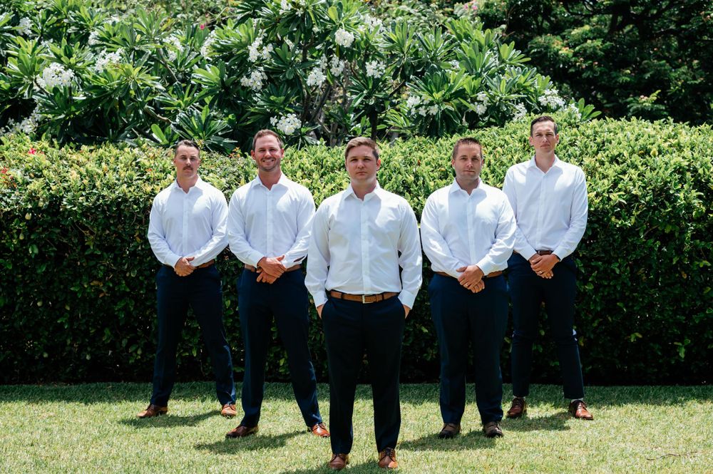 Wedding party in white shirts and dark pants posing together on sunny lawn.