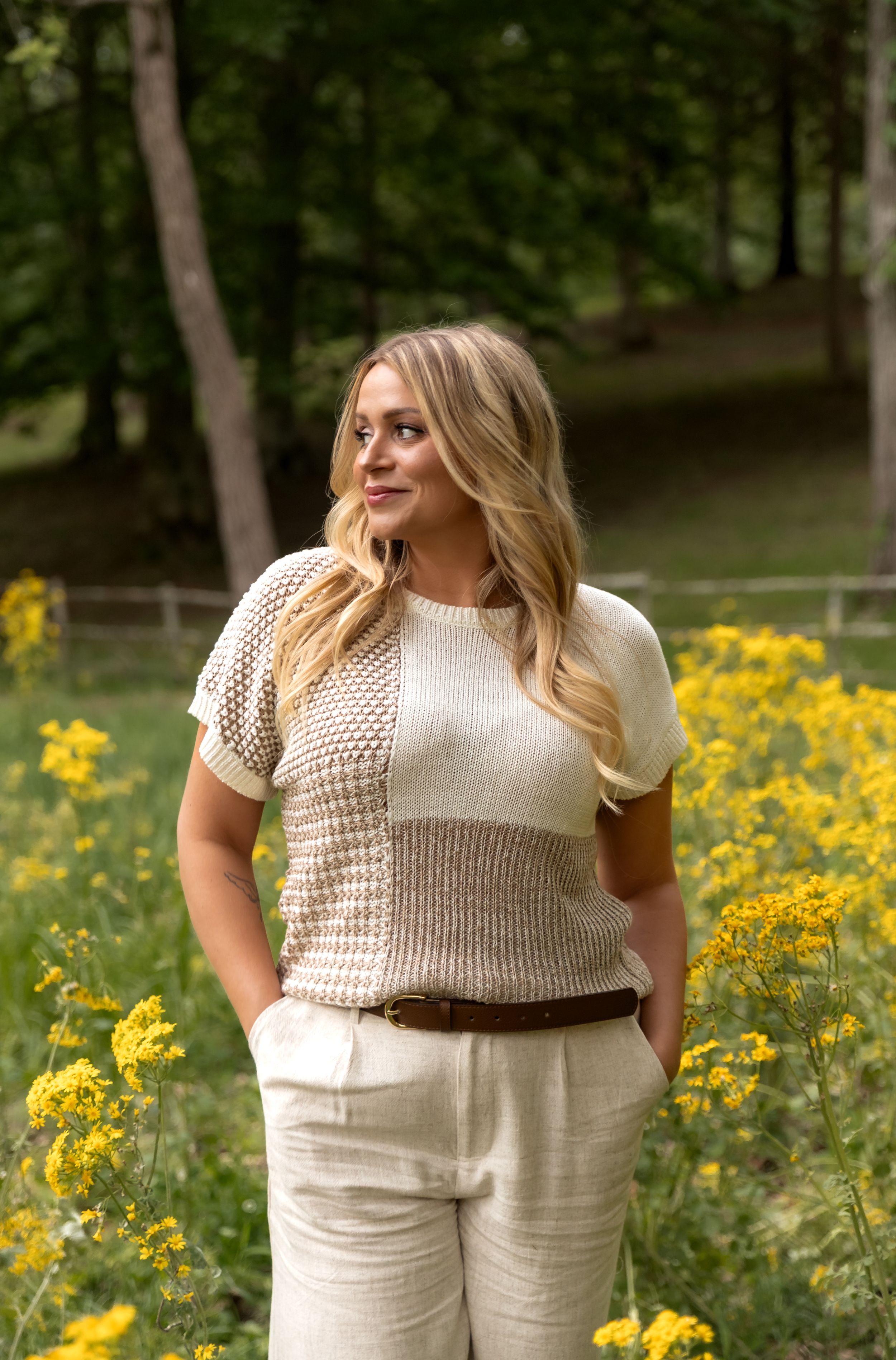 A fashion photoshoot in a meadow with yellow wildflowers featuring a cream colored knit top and white pants outfit.