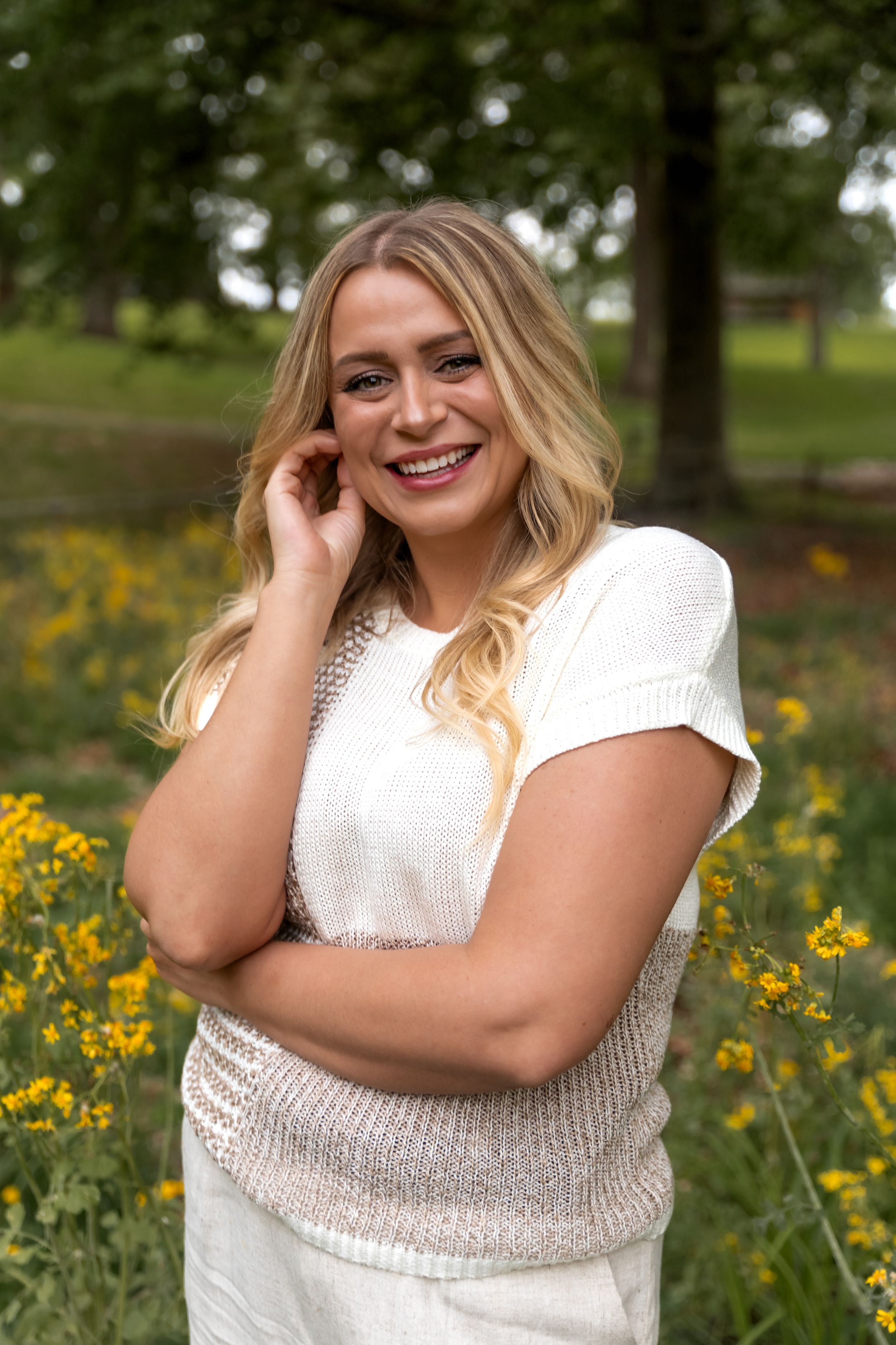 A woman in a white sweater poses cheerfully in a field of yellow wildflowers on a sunny day.