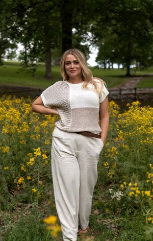 A person wearing a cream-colored knit sweater and white pants stands among blooming yellow wildflowers in a park setting.