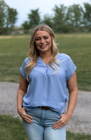 Someone wearing a light blue v-neck blouse and jeans poses outdoors with hands in pockets against a natural background.