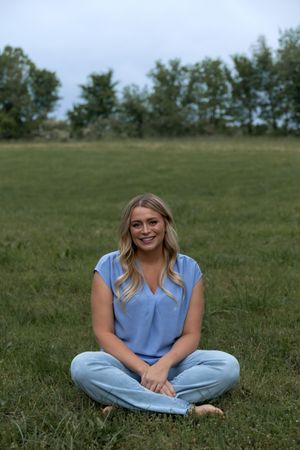 A person wearing a light blue shirt sits cross-legged on green grass in a summer field.