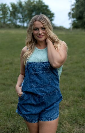 A person in denim overalls poses playfully on a green grassy slope.