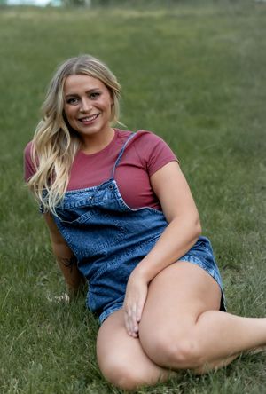 Person in denim overalls and mauve shirt relaxes on grassy field during outdoor portrait session.