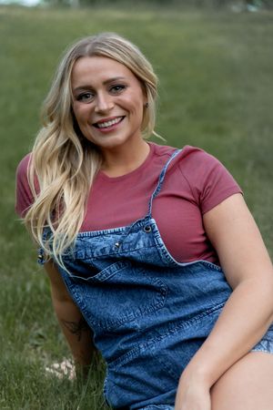 Person wearing denim overalls and pink t-shirt poses outdoors against natural green background.