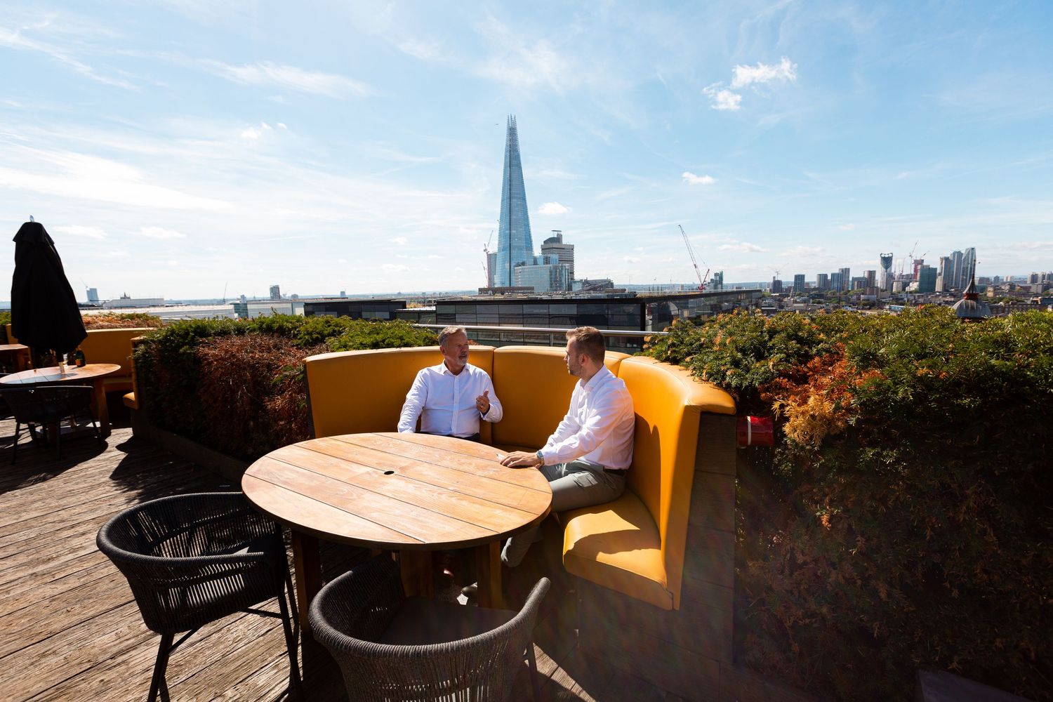 Two people enjoy drinks on a rooftop terrace overlooking London's skyline with The Shard visible in the distance.