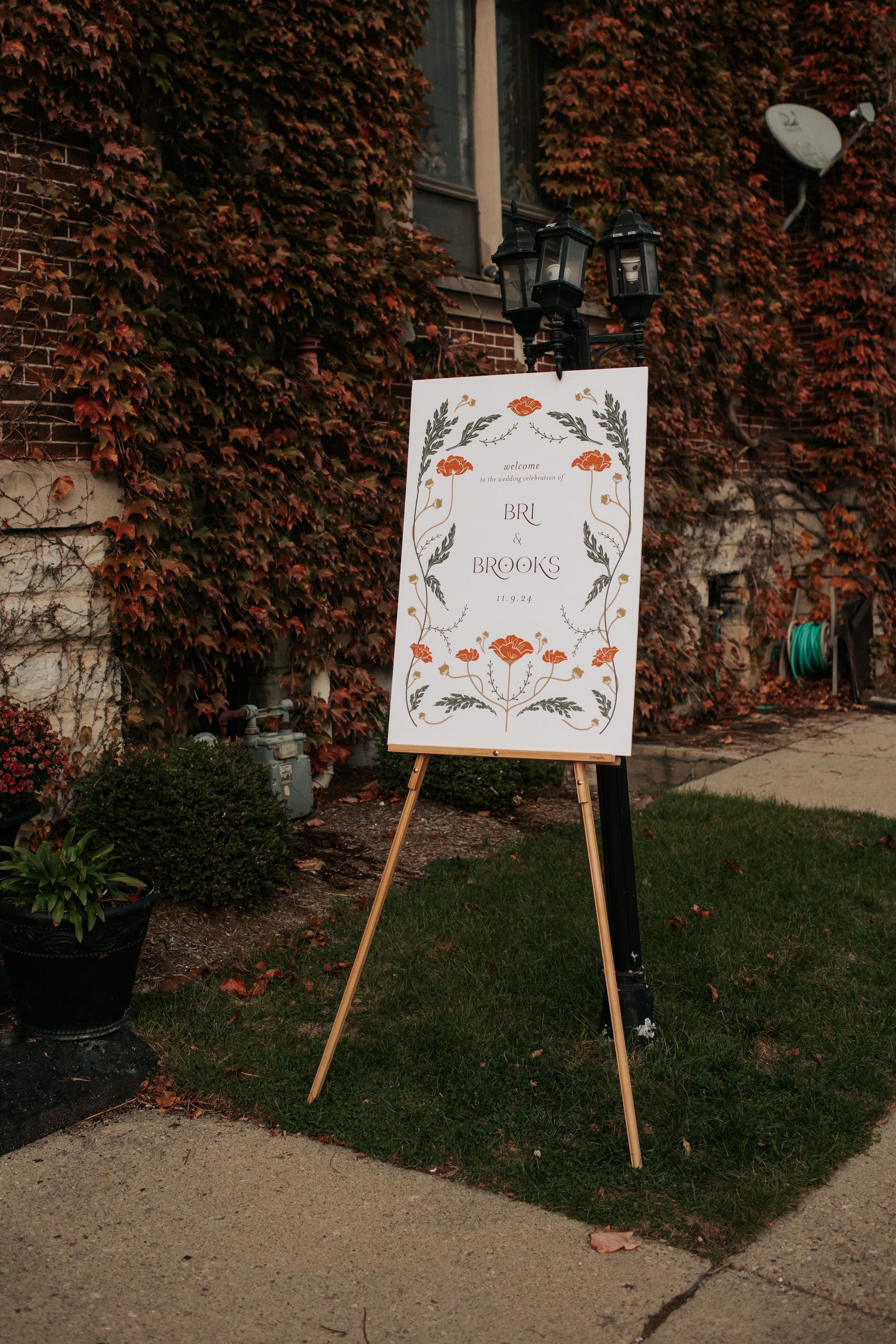 A white wedding welcome sign with floral design stands on a wooden easel against a brick wall covered in red ivy.