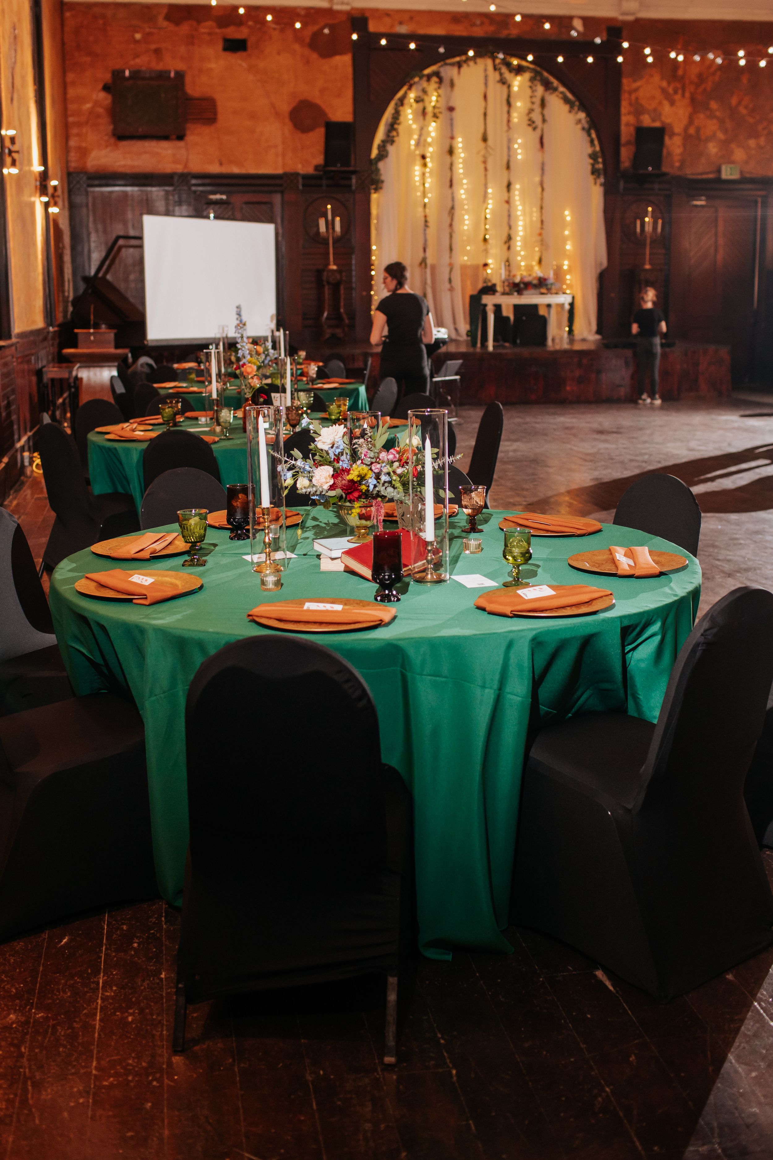 Wedding reception tables decorated with emerald green tablecloths, orange charger plates and black chair covers in a rustic venue.