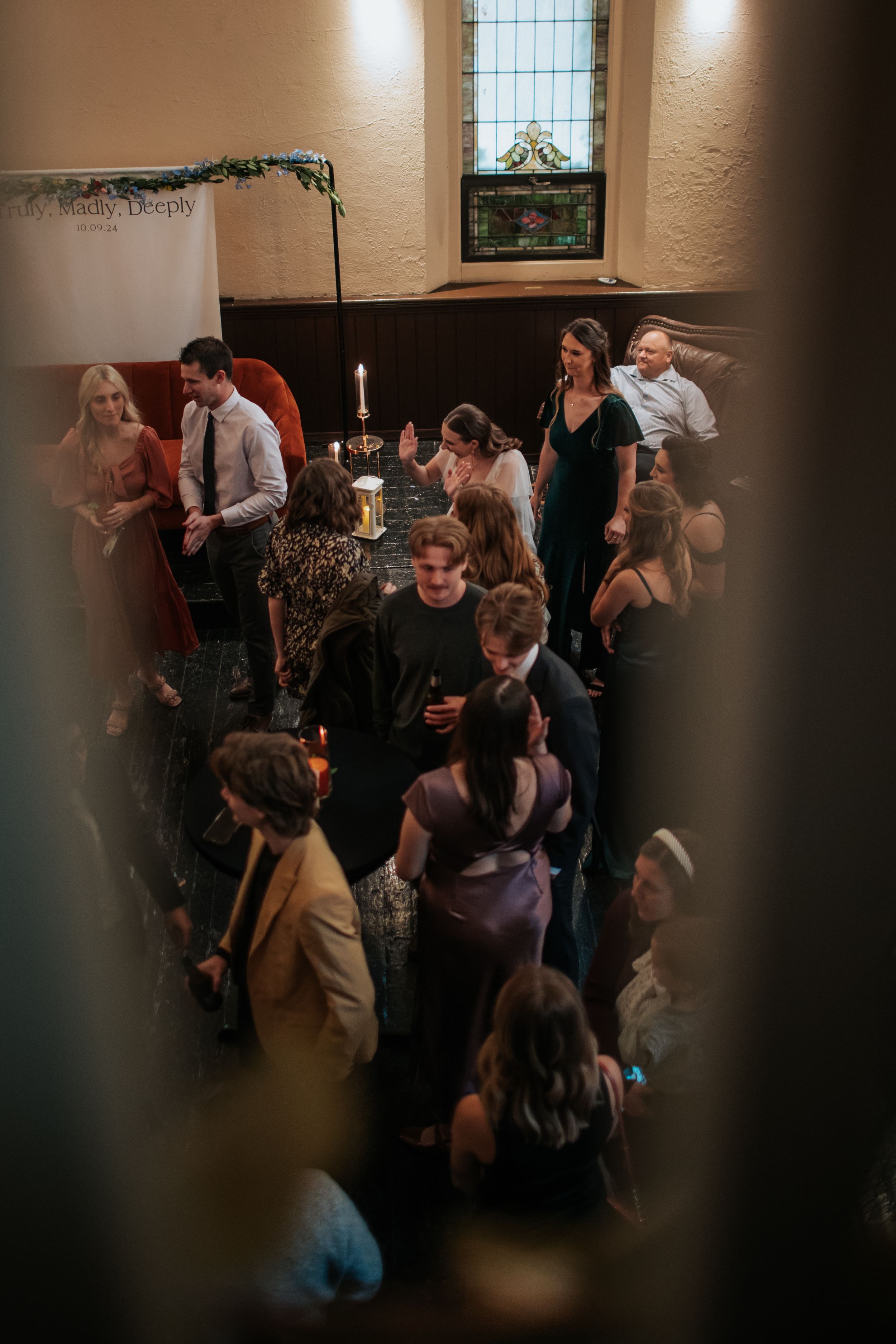 A dimly lit gathering of people socializing at an indoor event with a stained glass window and candlelight in the background.