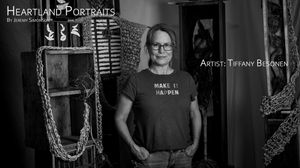 A black and white portrait of an artist wearing glasses and a dark t-shirt, standing in front of a decorated wall.
