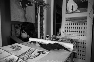 Black and white photo of a messy workspace with art supplies, papers, and drawing materials scattered across a desk.
