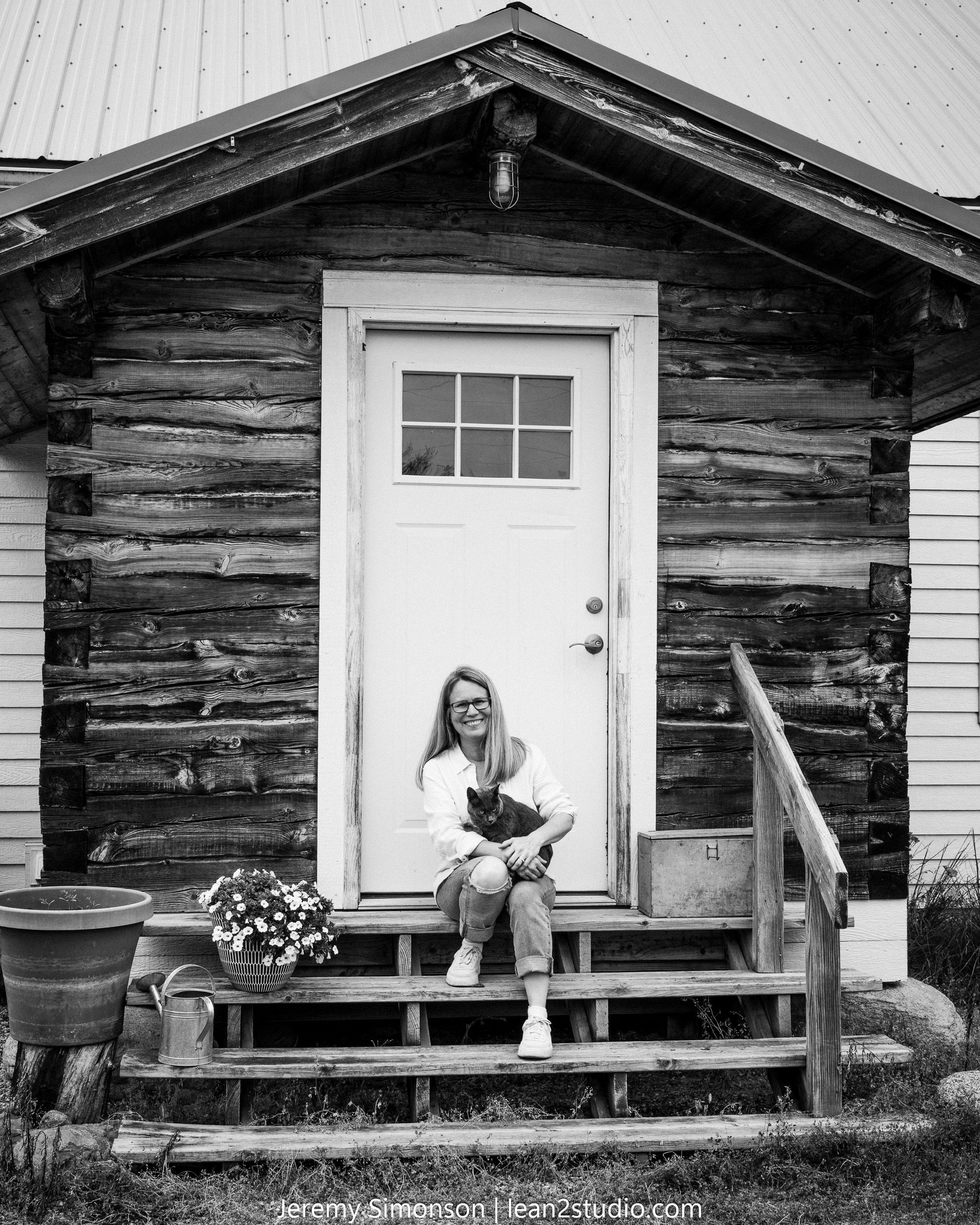Black and white photo of a rustic log cabin with a white door, wooden steps, and a potted plant on the porch.