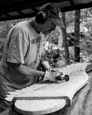 A craftsman in a baseball cap carefully shapes a wooden surfboard with hand tools in black and white photography.