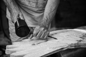 Hands use a mallet and chisel to carve details into a weathered piece of wood in black and white.