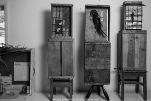 Three vintage wooden bird cages mounted on stands display exotic parrots in a black and white photograph.