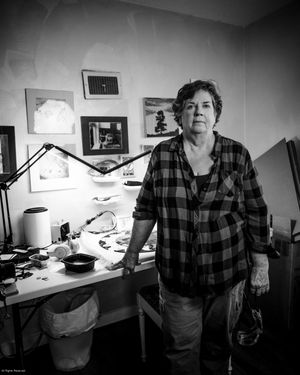 An artist stands in their workspace with tools and supplies on a desk in a black and white photograph.