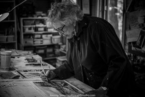 Black and white photo showing a person working at a desk with papers and documents scattered across the surface.