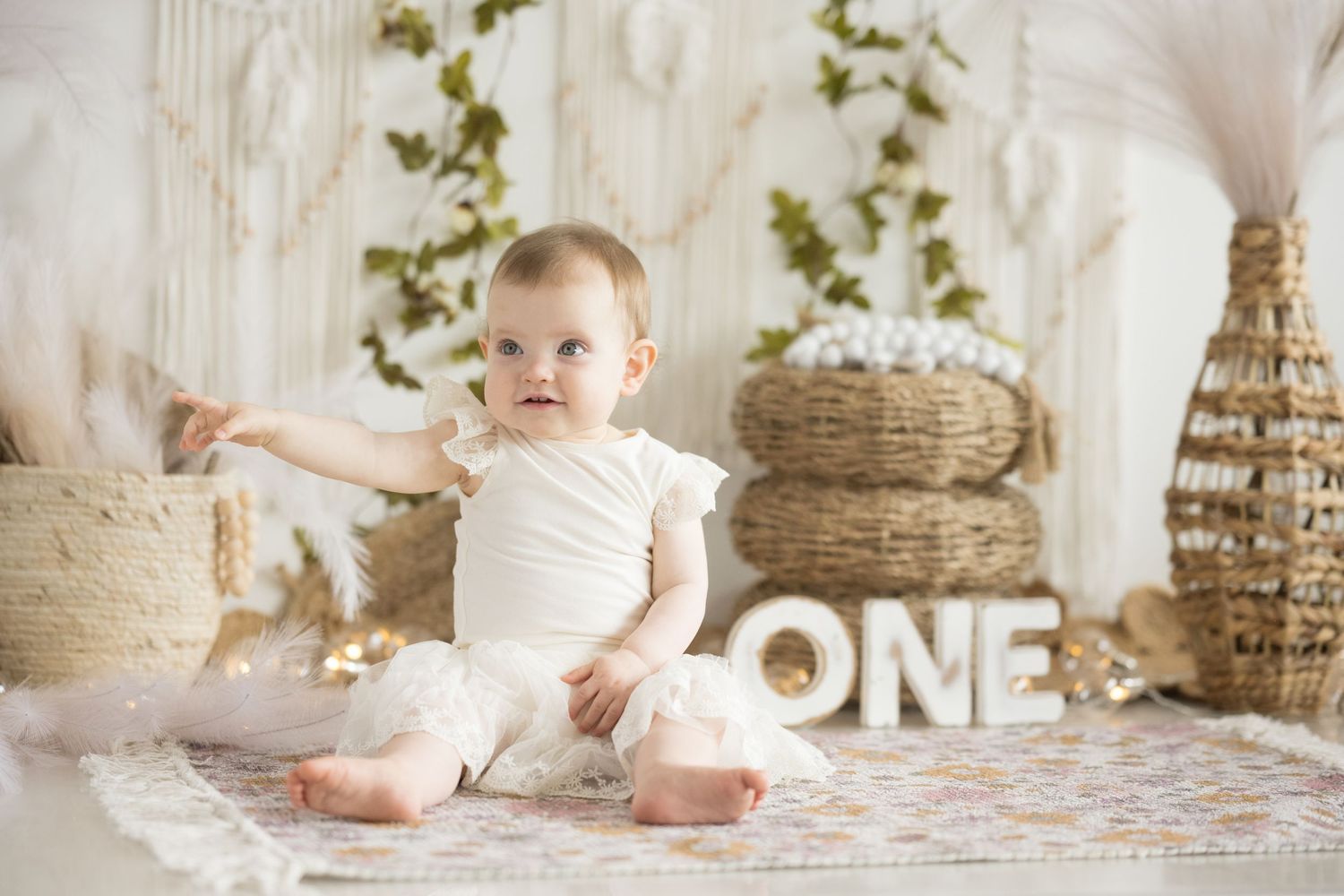 Baby in white outfit sitting by decorative ONE sign and natural baskets.