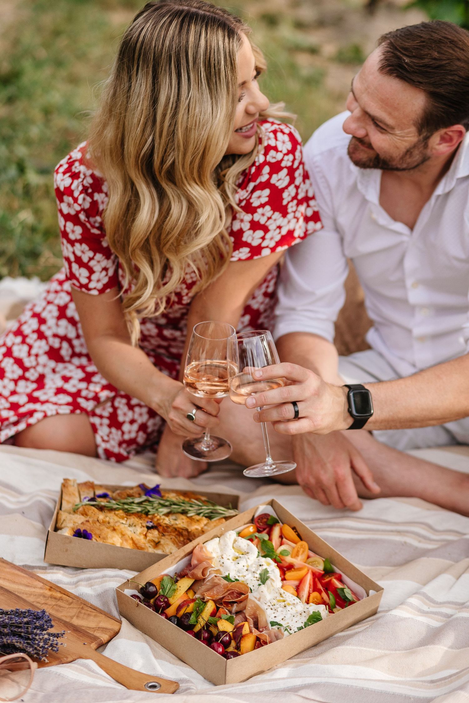 Couple's photoshoot enjoying romantic outdoor picnic with wine and charcuterie board on white blanket at Quails Gate winery in West Kelowna