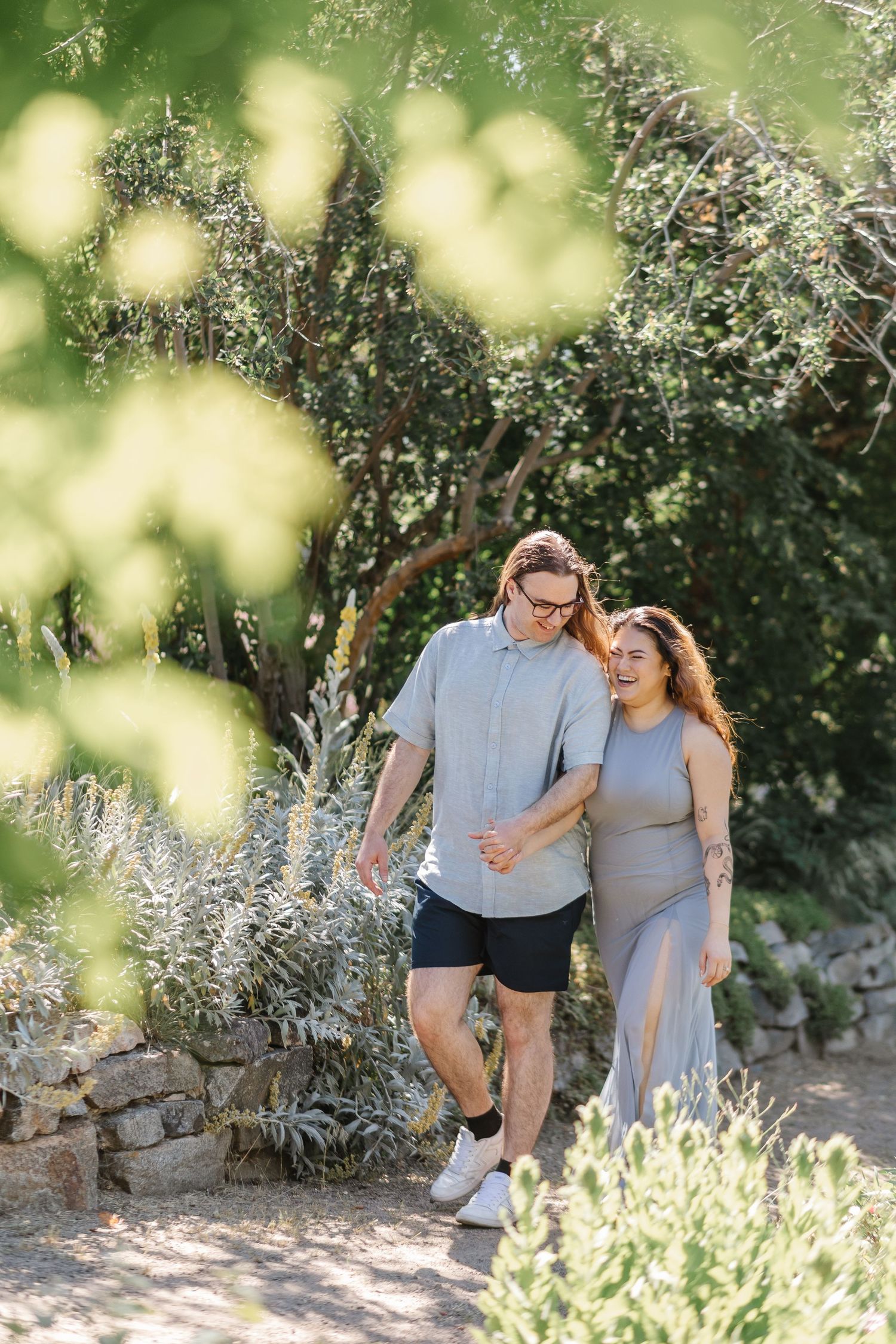 A couple walking along a path with sunlit bokeh effects at the Ornamental Gardens in Summerland, BC for engagement photos