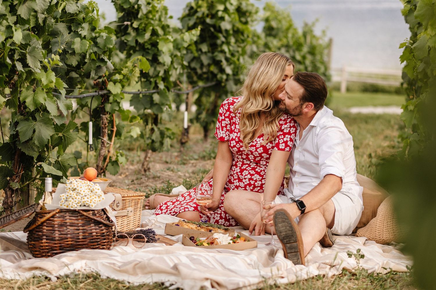 A couple enjoys a romantic picnic among lush green grapevines in a sunny vineyard setting at Quails Gate Winery for an engagement photoshoot