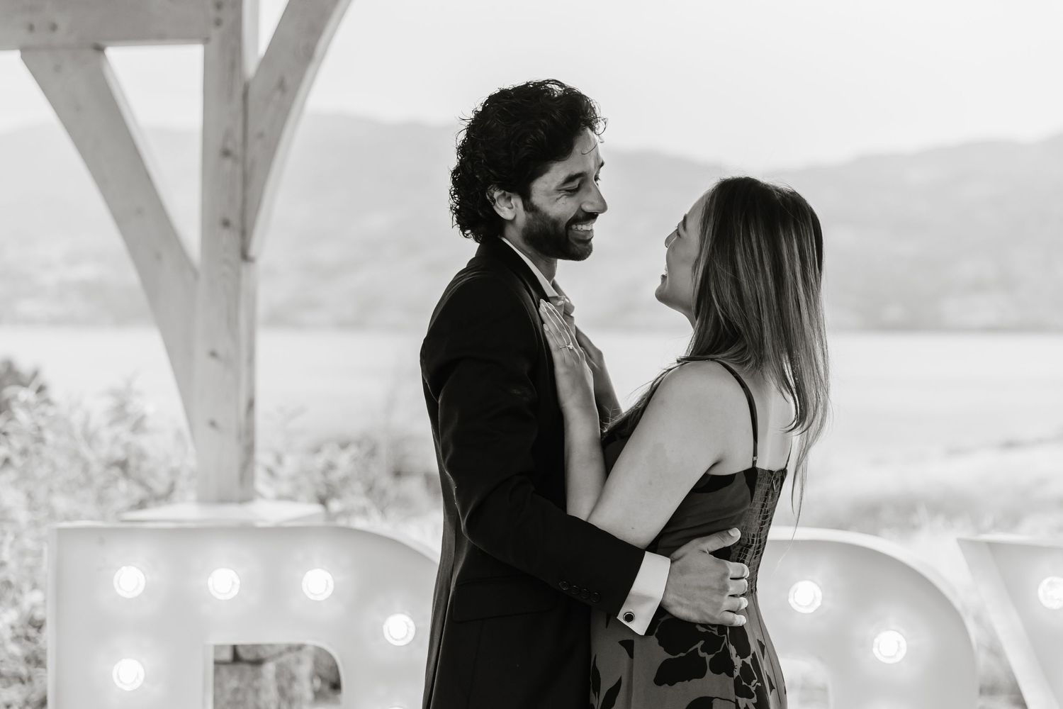 A romantic black and white photo of a couple embracing on a balcony with glowing lights at sanctuary gardens in West Kelowna BC for proposal