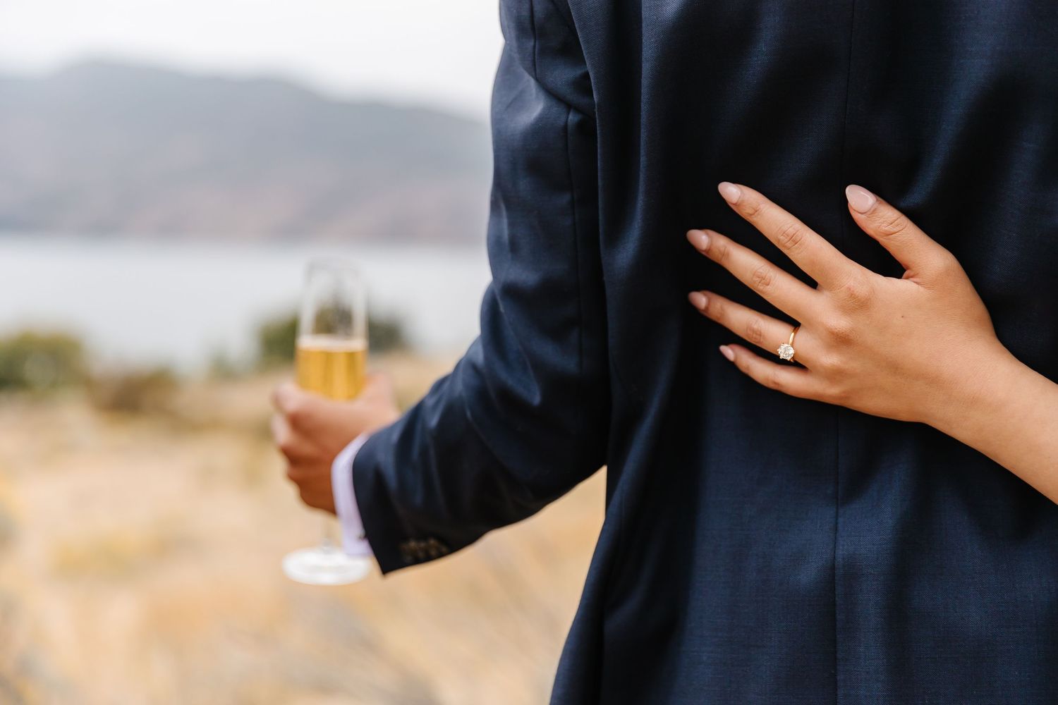 Close up of hands holding champagne glass during outdoor engagement celebration with scenic view in background of Okanagan lake in Kelowna