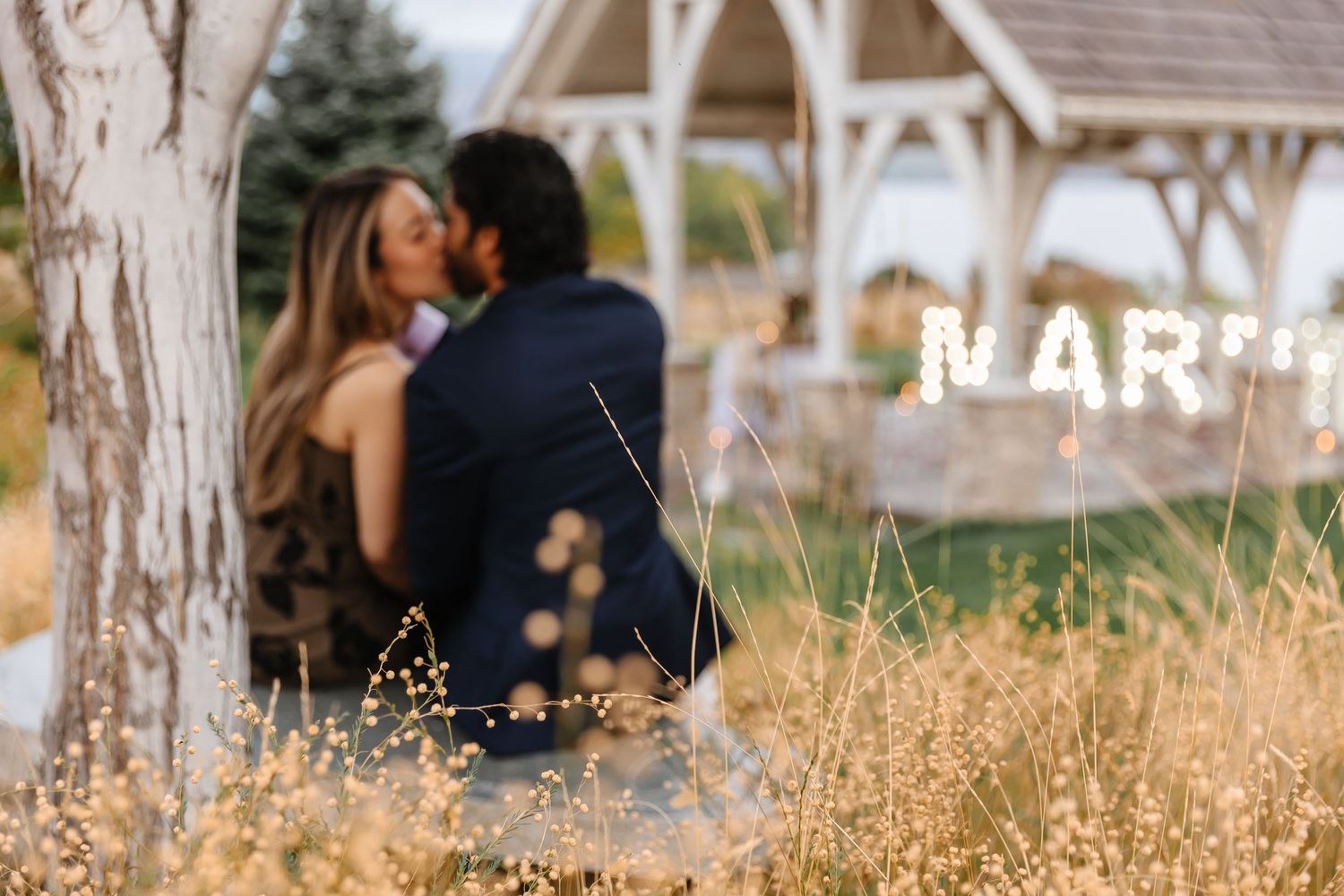Romantic couple sits in golden autumn field near white gazebo during sunset engagement photo session at sanctuary gardens in West Kelowna