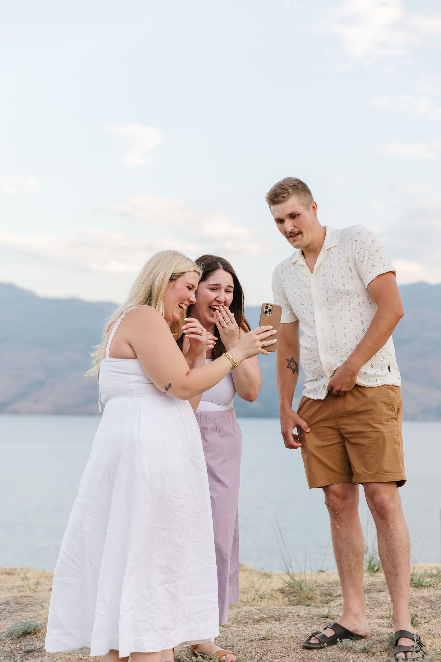A couple shares a joyful moment together while standing on a scenic hill with a view of Okanagan Lake after surprise proposal photoshoot.