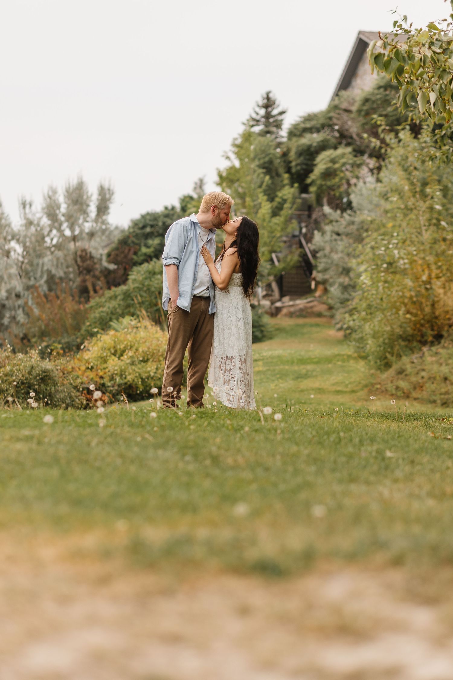 A romantic couple has outdoor engagement photos taken in a natural setting with soft sunlight filtering through green foliage in Kelowna, BC