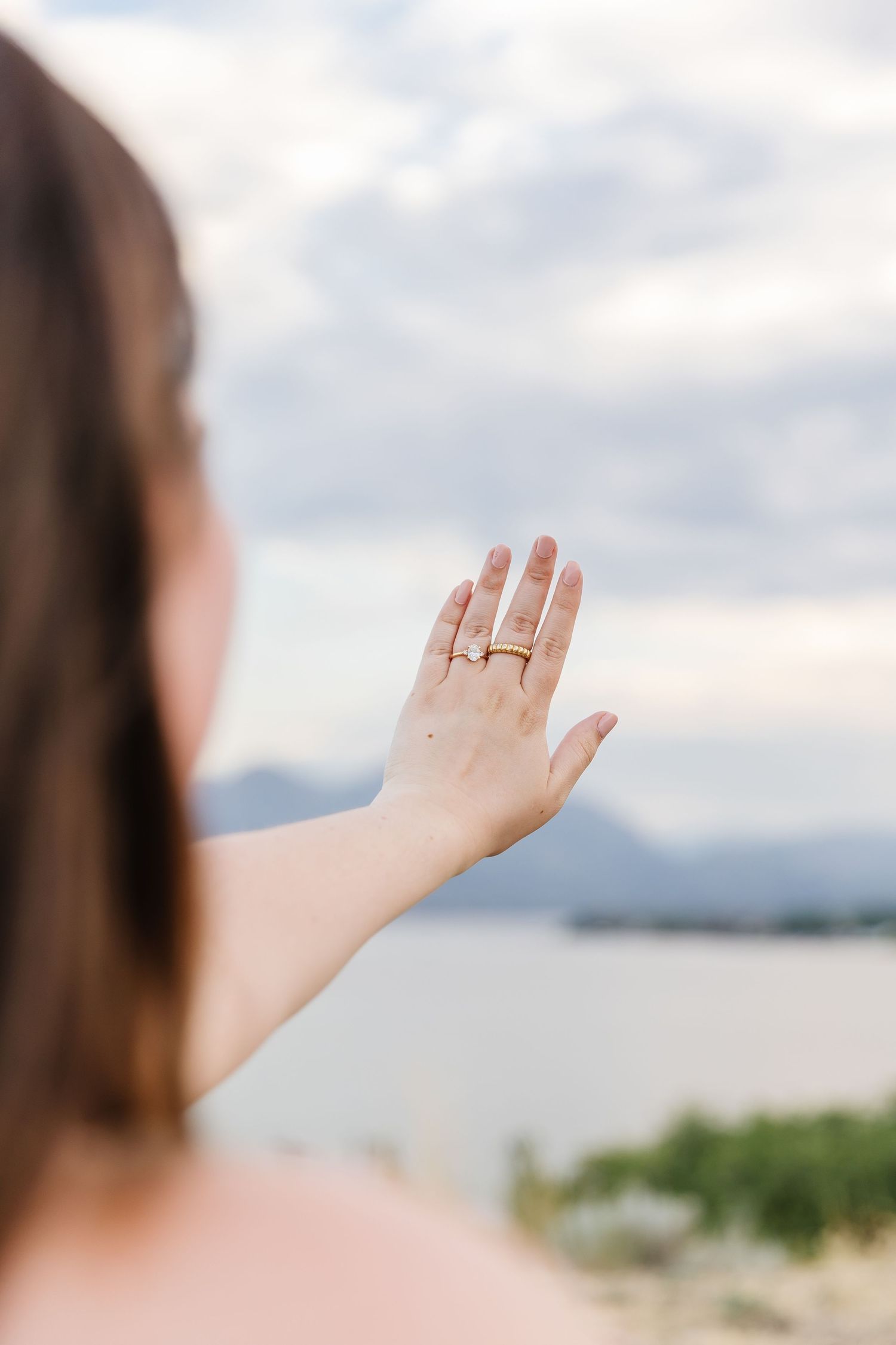 A hand wearing a new engagement ring is held up against a scenic mountain and Okanagan Lake during a surprise proposal photoshoot in Kelowna