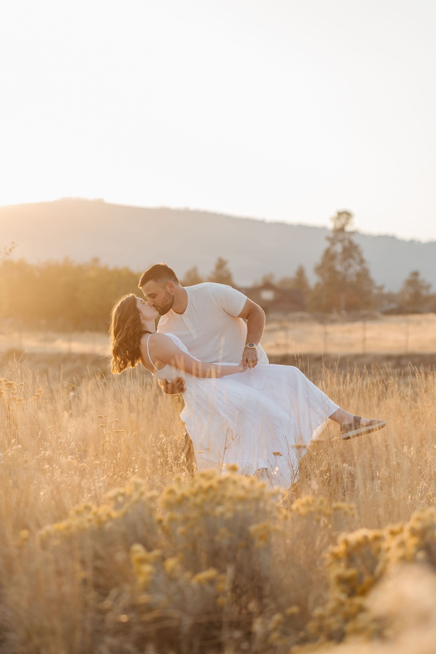 A romantic couple shares a tender dip kiss in a golden sunlit field at sunset in West Kelowna, BC for an engagement photoshoot.