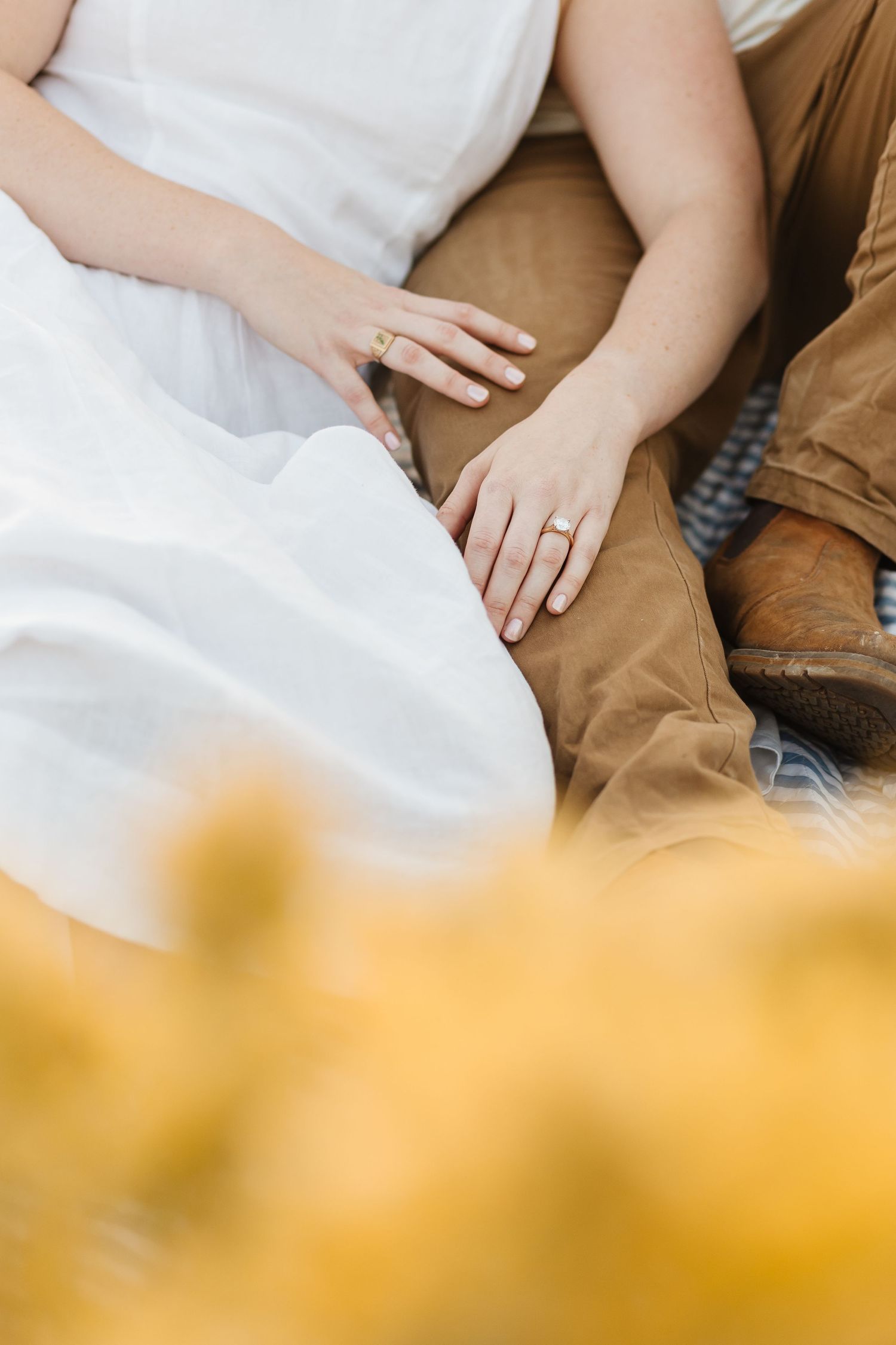 Close-up of hands with engagement ring resting on brown fabric, with soft yellow bokeh in foreground during engagement photoshoot in Kelowna