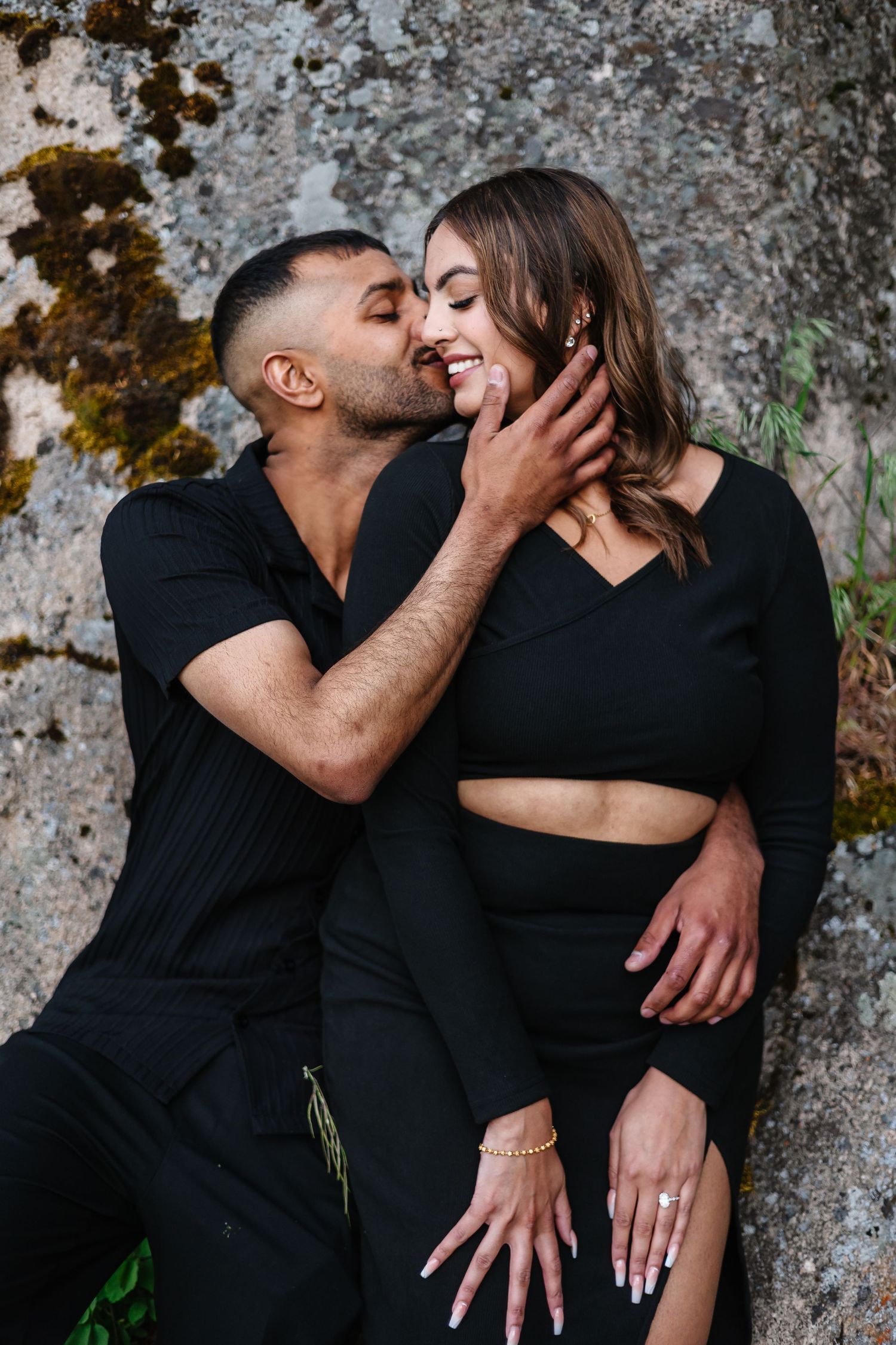A romantic couple in black outfits sharing an intimate moment against a textured stone wall at sparkling hills for a surprise proposal photo